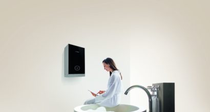 Woman wearing white bathrobe sitting on edge of modern bathtub and using digital tablet